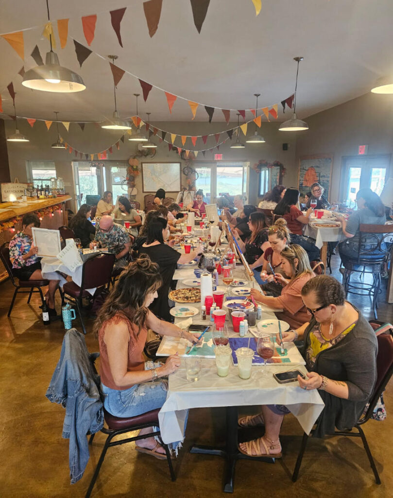 group of people sitting at long table painting at an event with bunting on the ceiling